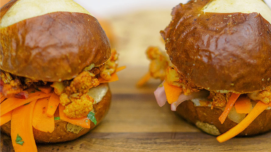 Two BBQ Cauliflower Sliders with pretzel buns sitting on a wooden board