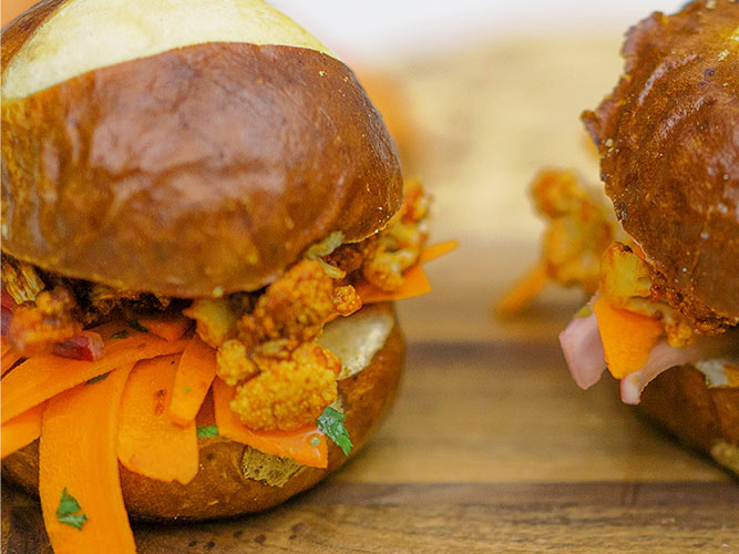 Two BBQ Cauliflower Sliders with pretzel buns sitting on a wooden board