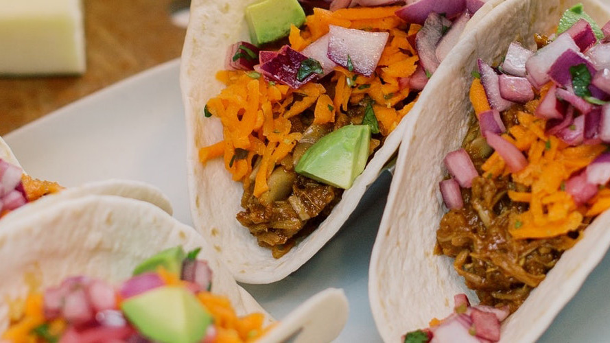 BBQ Jackfruit Tacos on soft shell tortillas standing up so you can see what's inside- avocado, cheese, red onions, and meat