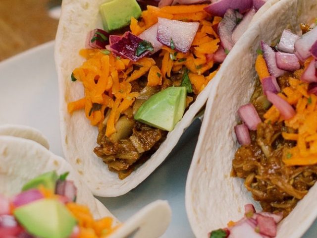 BBQ Jackfruit Tacos on soft shell tortillas standing up so you can see what's inside- avocado, cheese, red onions, and meat