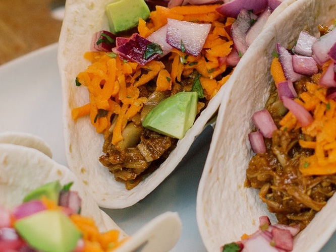 BBQ Jackfruit Tacos on soft shell tortillas standing up so you can see what's inside- avocado, cheese, red onions, and meat