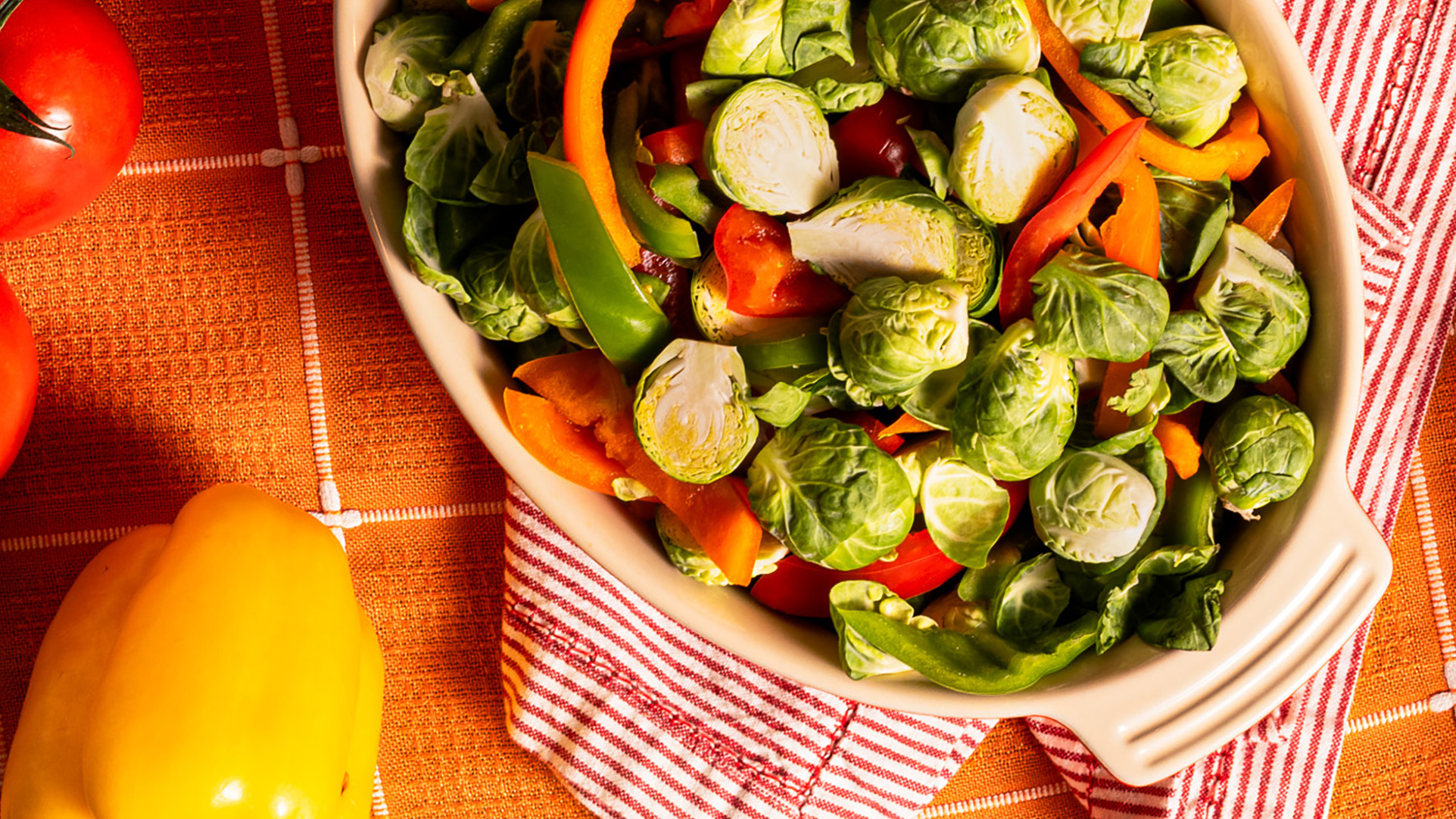 Dorothy Lynch Brussel Sprouts in a baking dish with splashed of orange, yellow, and red in the table cloth, hand towels, and peppers surrounding the dish