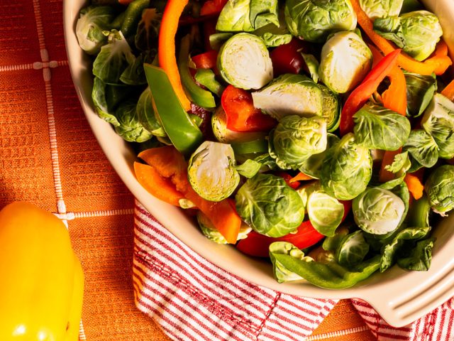 Dorothy Lynch Brussel Sprouts in a baking dish with splashed of orange, yellow, and red in the table cloth, hand towels, and peppers surrounding the dish