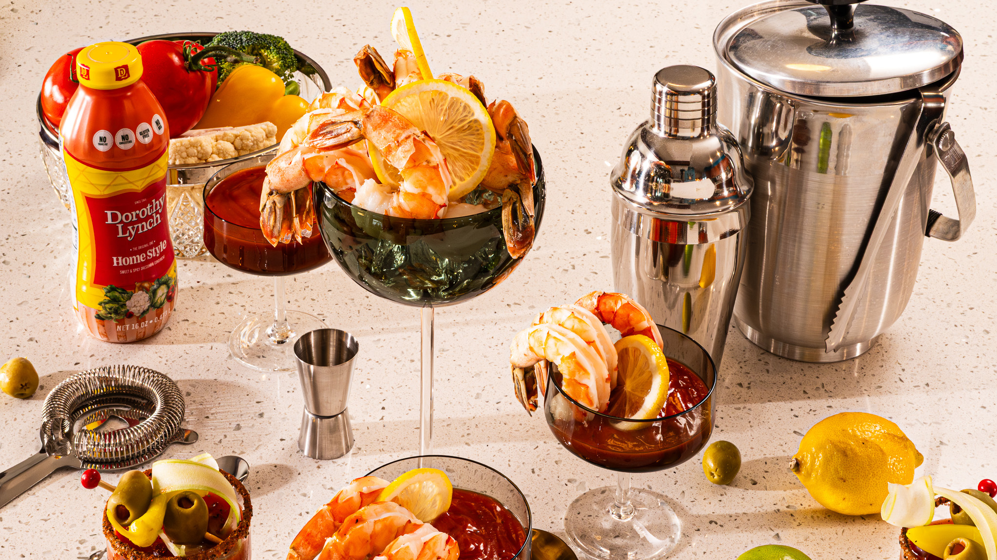 Dorothy Lynch Dressing Shrimp Cocktail Sauce in fancy glass with lots of cocktail equipment , veggies, and Dorothy Lynch Dressing scattered around