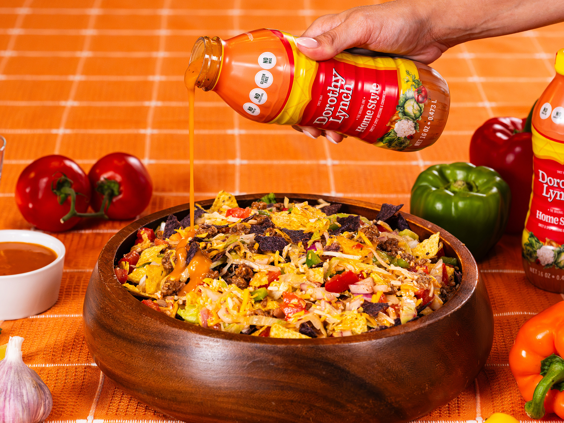 Hand pouring Dorothy Lynch Dressing on top of a Meatless Taco Salad Recipe in a wooden bowl on a orange checkered table cloth with other veggies surrounding the place setting