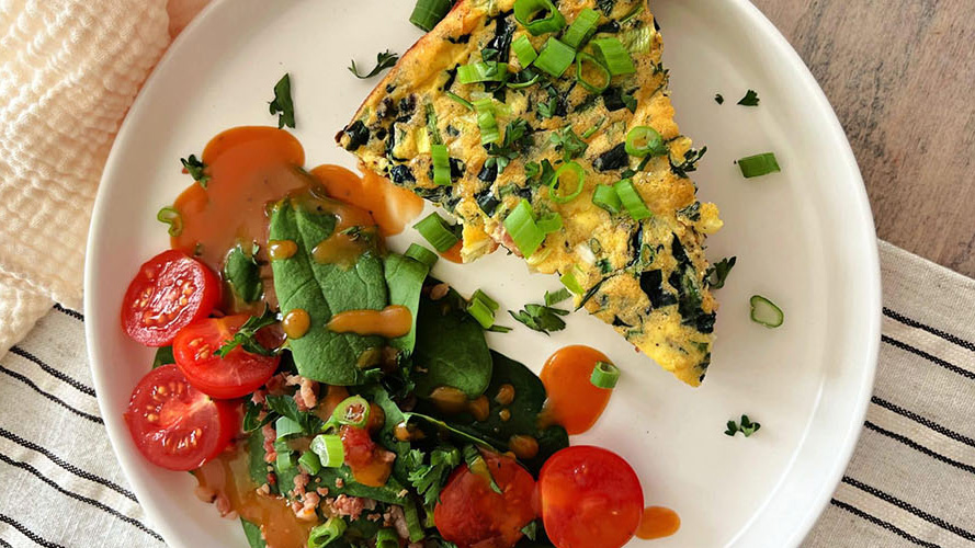 Triangle slice of Dorothy Lynch Bacon Potato Frittata on a white plate with a salad, including spinach, tomato, and Dorothy Lynch, laying next to it