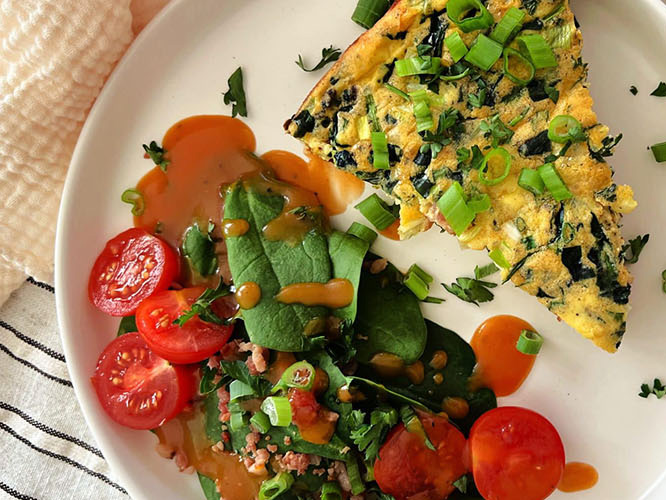 Triangle slice of Dorothy Lynch Bacon Potato Frittata on a white plate with a salad, including spinach, tomato, and Dorothy Lynch, laying next to it
