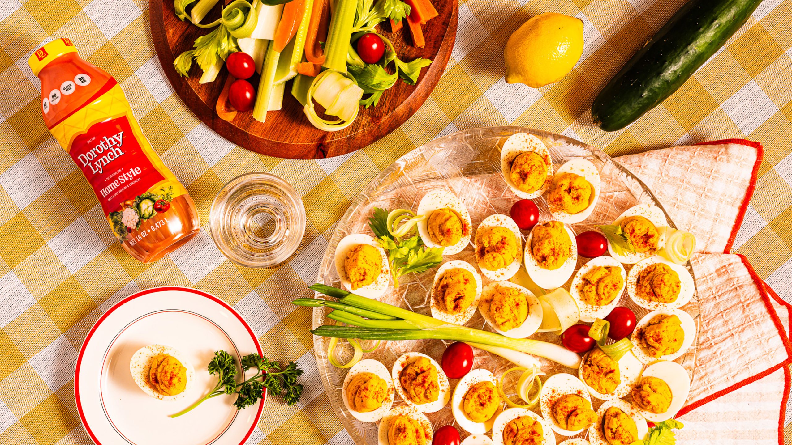 Dorothy Lynch Deviled Eggs on a clear circular platter. Yellow checkered table cloth underneath with some veggies and a Dorothy Lynch Dressing bottle laying beside the tray