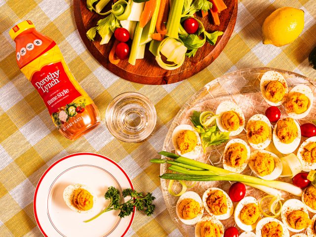 Dorothy Lynch Deviled Eggs on a clear circular platter. Yellow checkered table cloth underneath with some veggies and a Dorothy Lynch Dressing bottle laying beside the tray