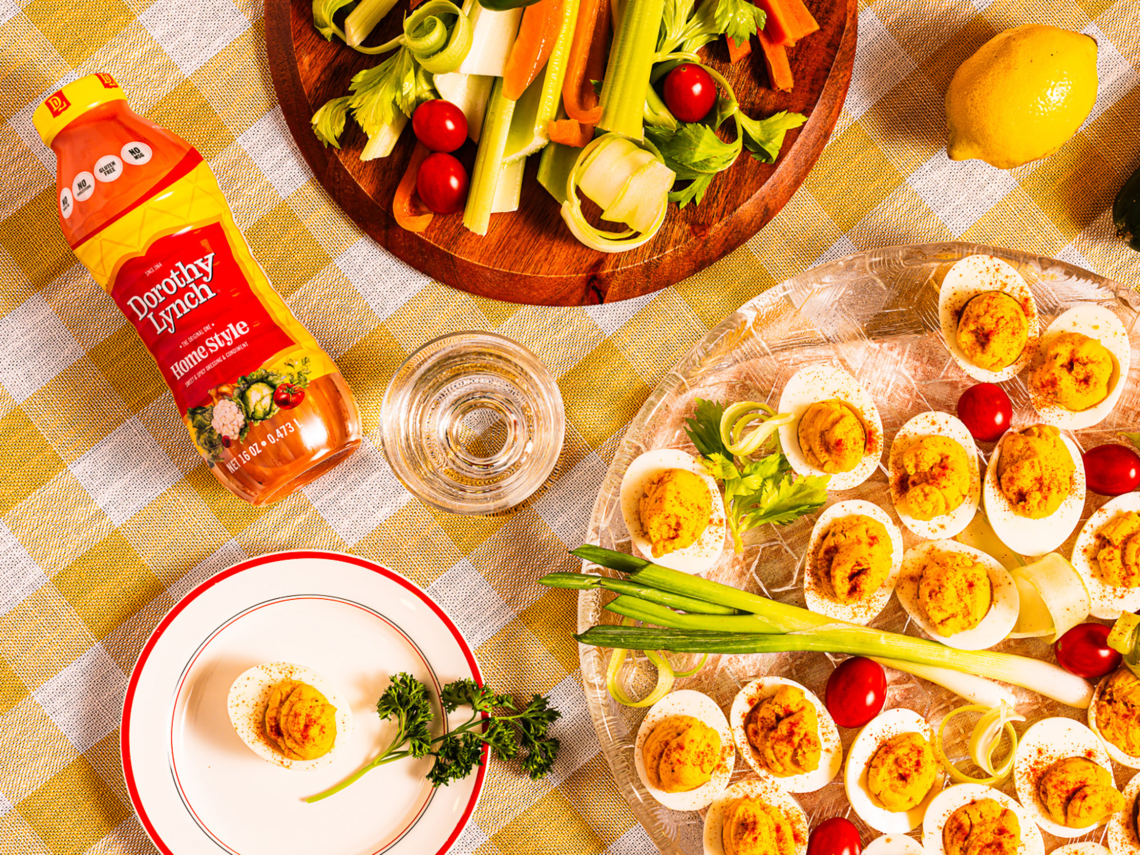 Dorothy Lynch Deviled Eggs on a clear circular platter. Yellow checkered table cloth underneath with some veggies and a Dorothy Lynch Dressing bottle laying beside the tray