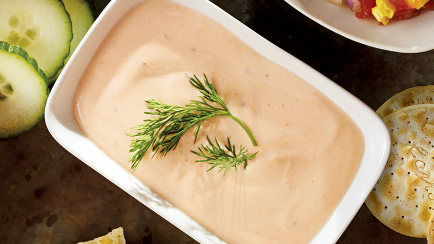 Dorothy’s Light & Lean Spicy Greek Dip in a white bowl with cucumbers and crackers laying beside on the table